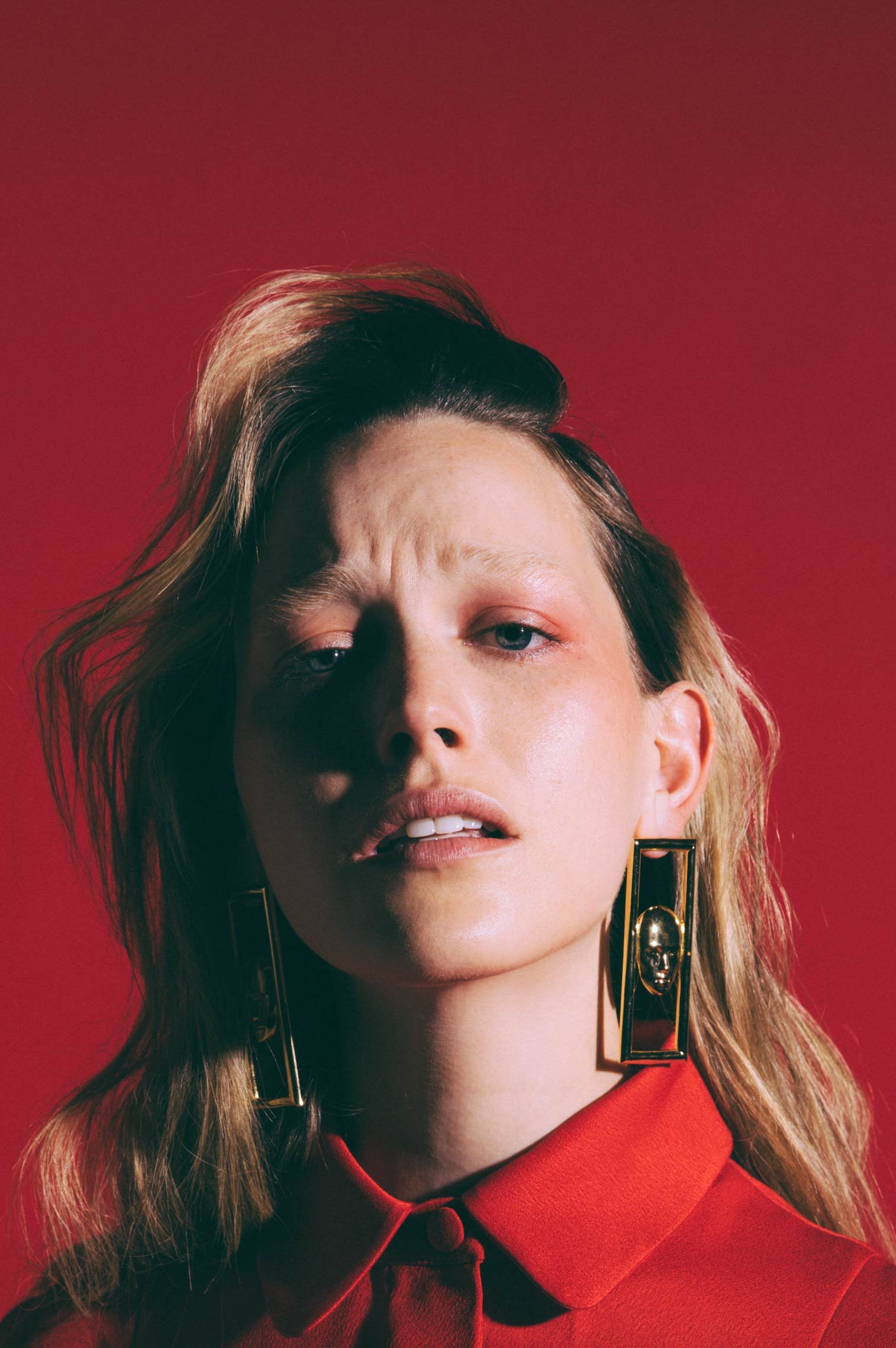 Blonde woman in a red collared shirt, looking up with a slight frown, wearing gold rectangle earrings, set against a red backdrop.