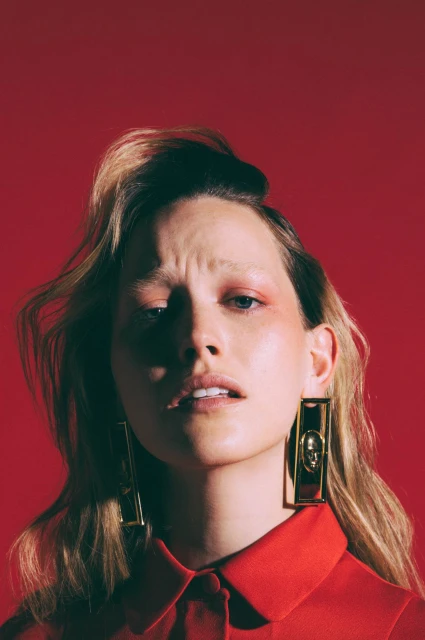 Blonde woman in a red collared shirt, looking up with a slight frown, wearing gold rectangle earrings, set against a red backdrop.