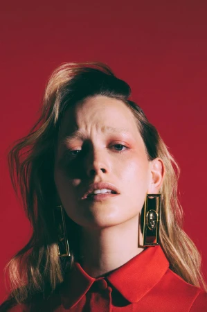 Blonde woman in a red collared shirt, looking up with a slight frown, wearing gold rectangle earrings, set against a red backdrop.