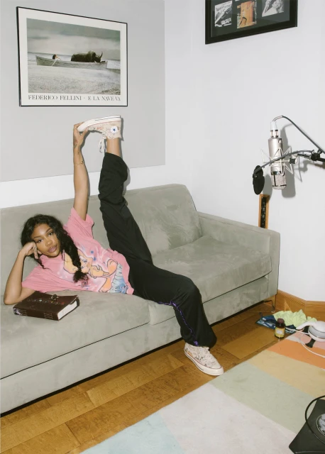 A young woman in black pants and a pink t-shirt poses on a gray couch with one leg raised high in an indoor setting with a microphone.