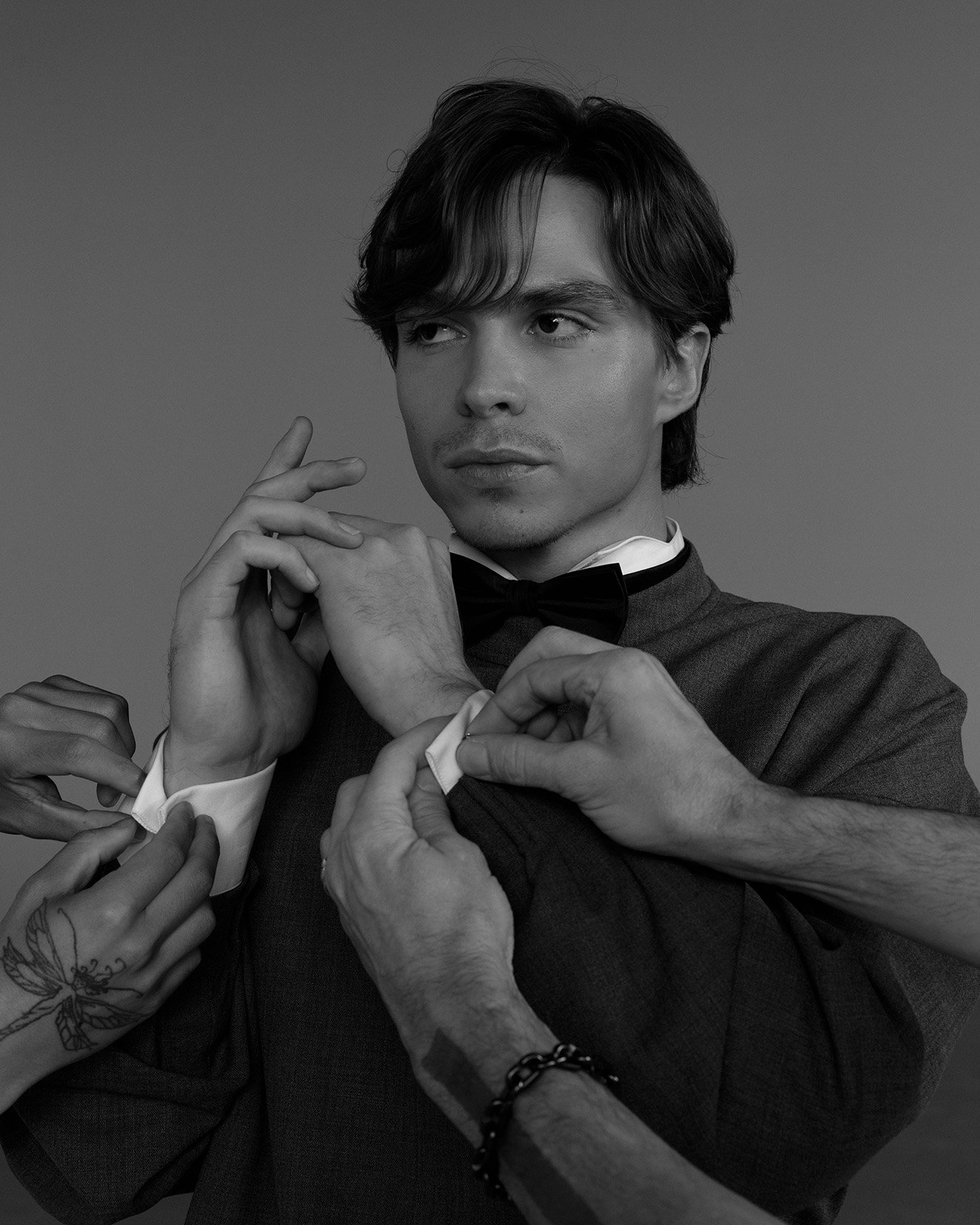 Black and white portrait of a young adult man in a suit with a bow tie, his cuffs being adjusted by multiple sets of hands.