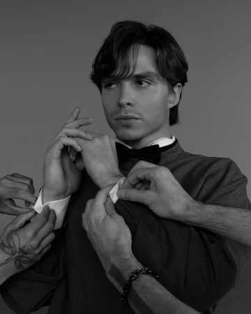 Black and white portrait of a young adult man in a suit with a bow tie, his cuffs being adjusted by multiple sets of hands.