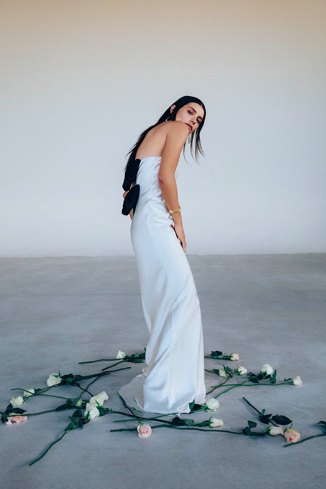 Woman in a white strapless dress stands amid a scattering of white roses on a grey floor against a stark white background.