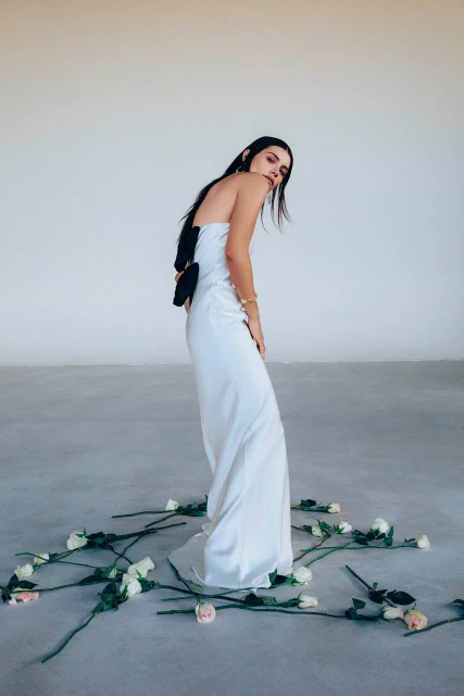 Woman in a white strapless dress stands amid a scattering of white roses on a grey floor against a stark white background.