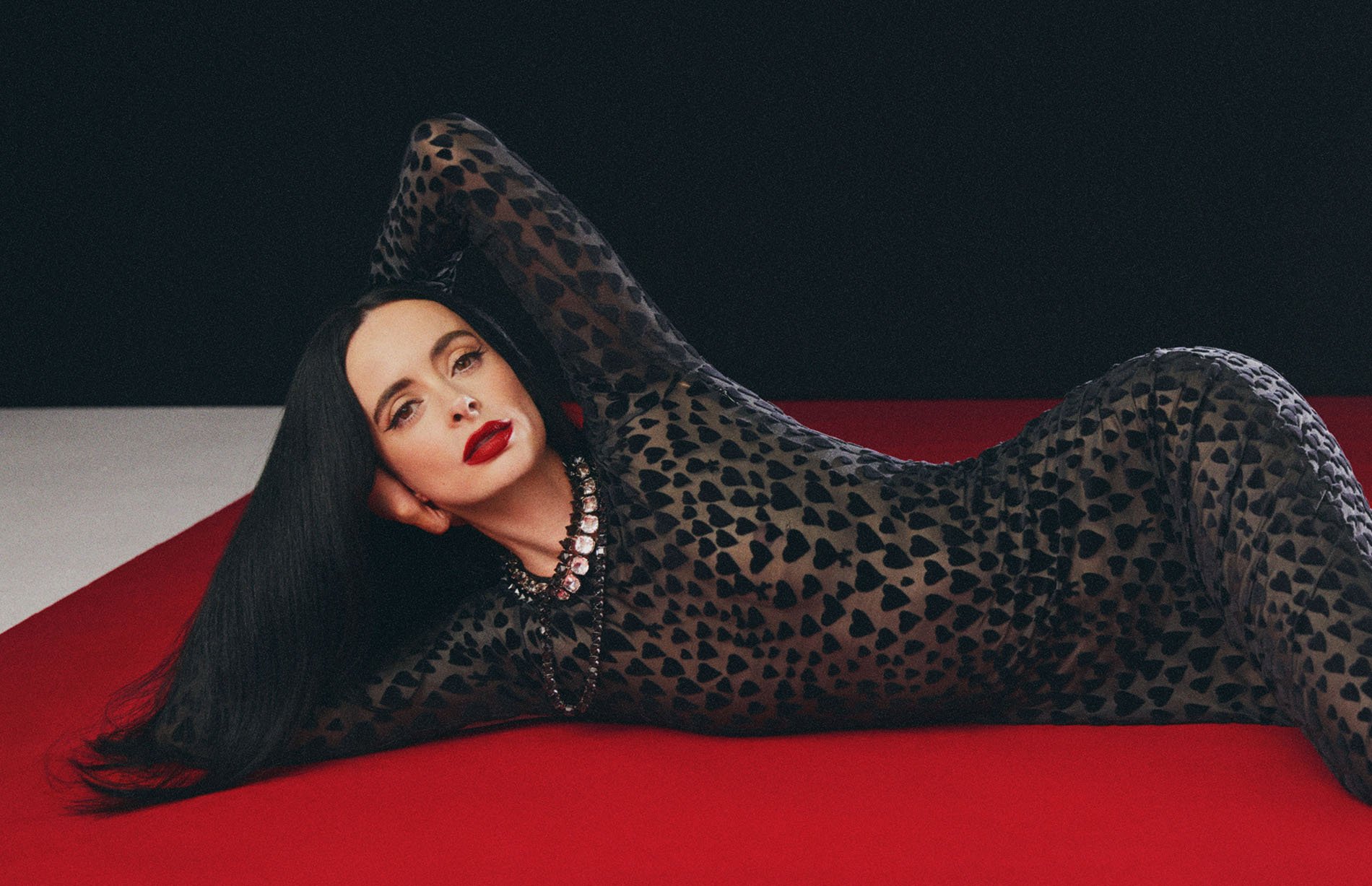 Woman in a sheer black heart-print dress lying on a red surface against a black and white backdrop, showcasing fashion and beauty.