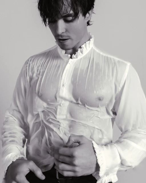 Monochrome portrait of a man in a wet, sheer white ruffled shirt, buttoning it closed, set against a light background.