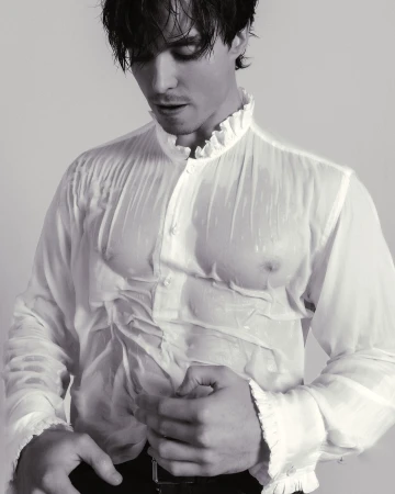 Monochrome portrait of a man in a wet, sheer white ruffled shirt, buttoning it closed, set against a light background.