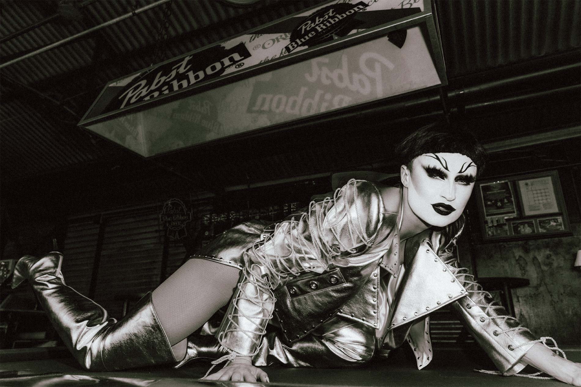 A person in silver clothing and boots poses on a green surface in a black-and-white photo in front of a Pabst Blue Ribbon sign.