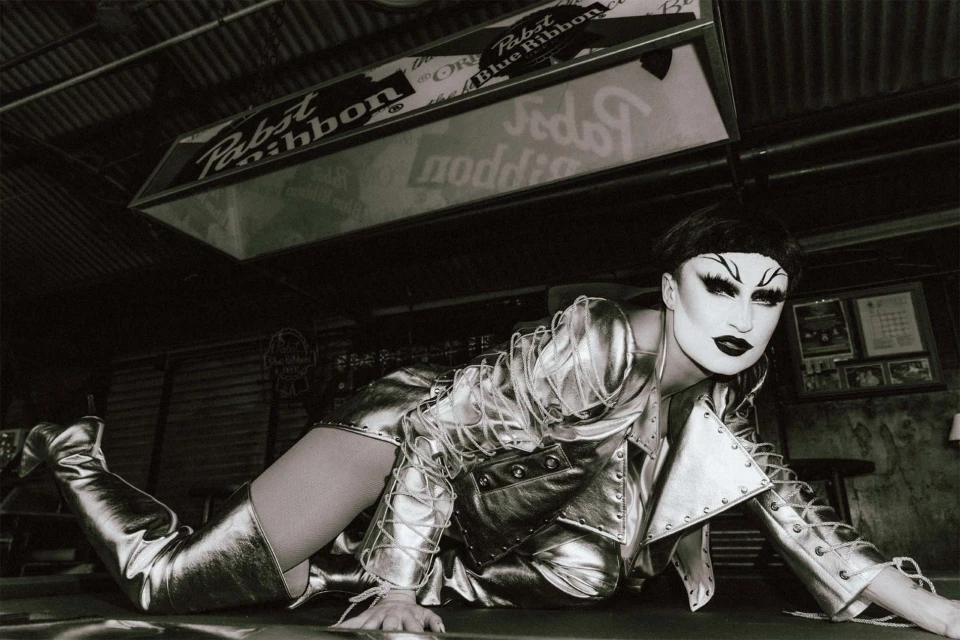 A person in silver clothing and boots poses on a green surface in a black-and-white photo in front of a Pabst Blue Ribbon sign.
