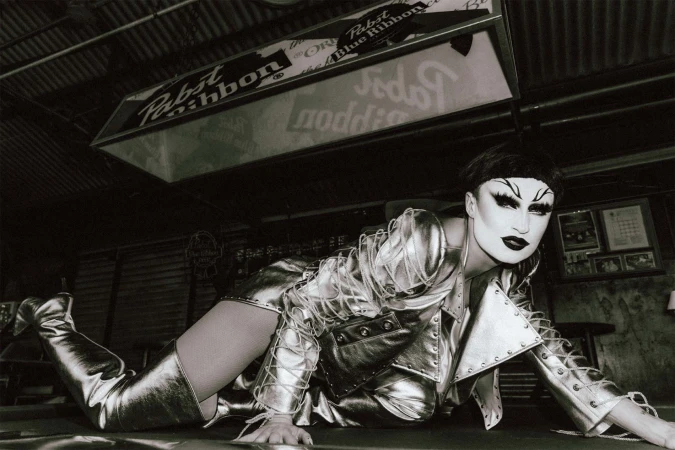 A person in silver clothing and boots poses on a green surface in a black-and-white photo in front of a Pabst Blue Ribbon sign.