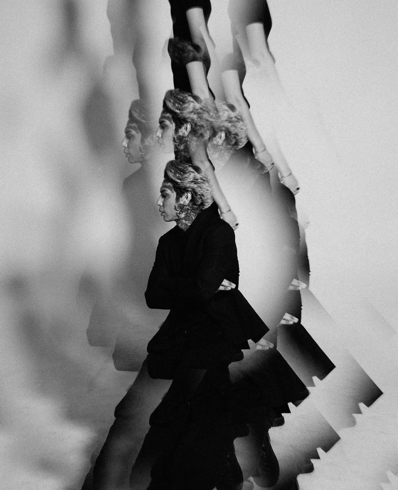Monochrome portrait of androgynous person with short curly hair in a dark jacket, duplicated and blurred against a neutral background.