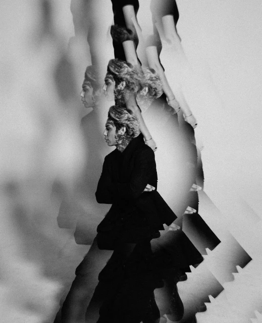Monochrome portrait of androgynous person with short curly hair in a dark jacket, duplicated and blurred against a neutral background.