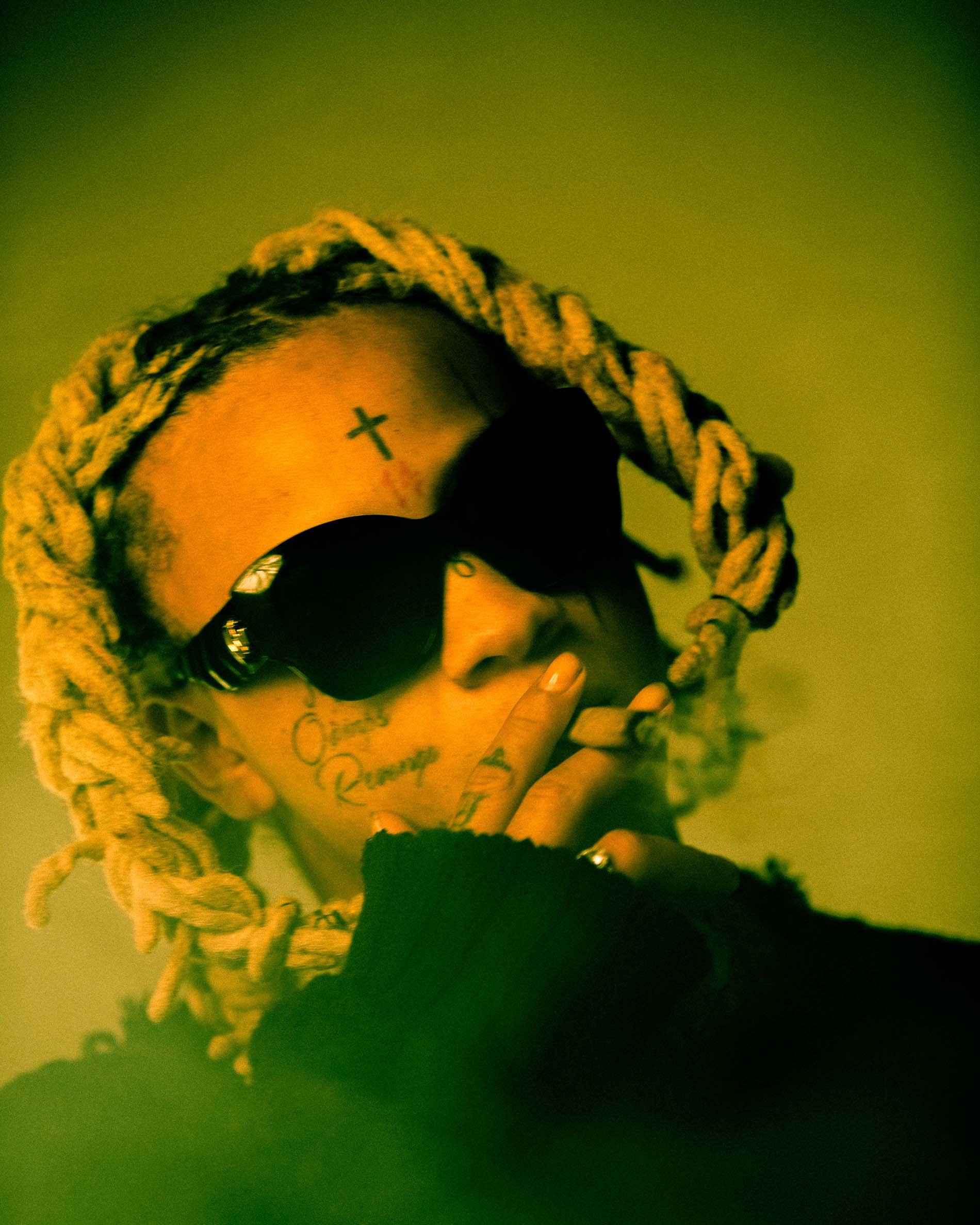 Portrait of a person with dreadlocks and sunglasses smoking, set against a soft green backdrop in a close-up composition.