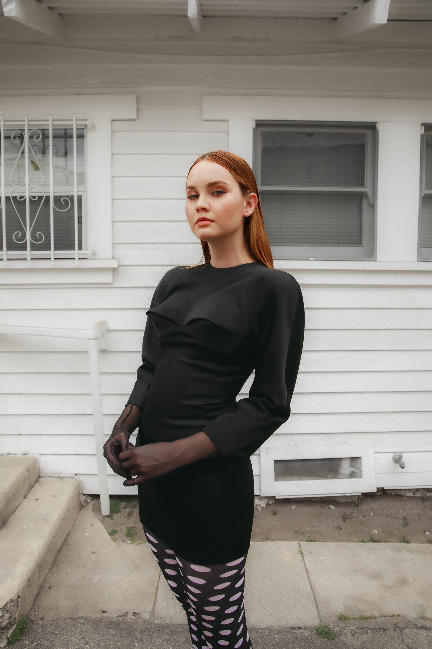 A woman with auburn hair poses in a black dress and sheer gloves in front of a white house, creating a chic and slightly edgy fashion portrait.