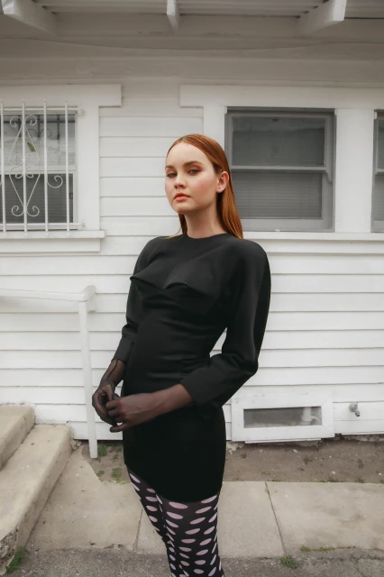 A woman with auburn hair poses in a black dress and sheer gloves in front of a white house, creating a chic and slightly edgy fashion portrait.