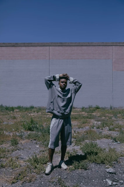 An adult Black male stands outdoors near a brick wall wearing a grey sweatshirt and shorts with his hands behind his head.