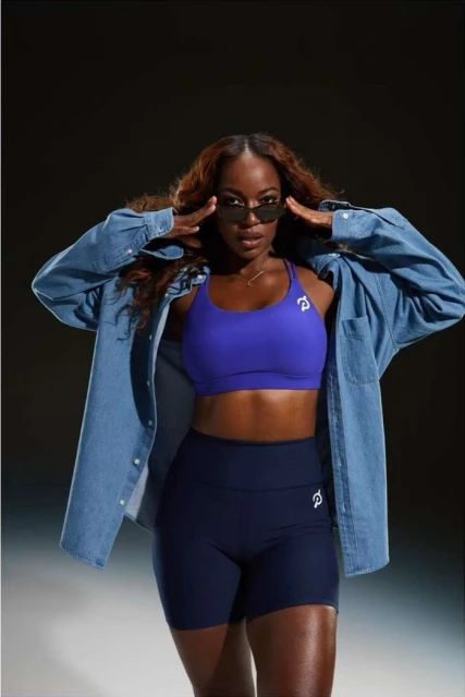 A Black female fitness model poses in a sports bra and shorts with a denim shirt, wearing sunglasses in a studio with dark background.