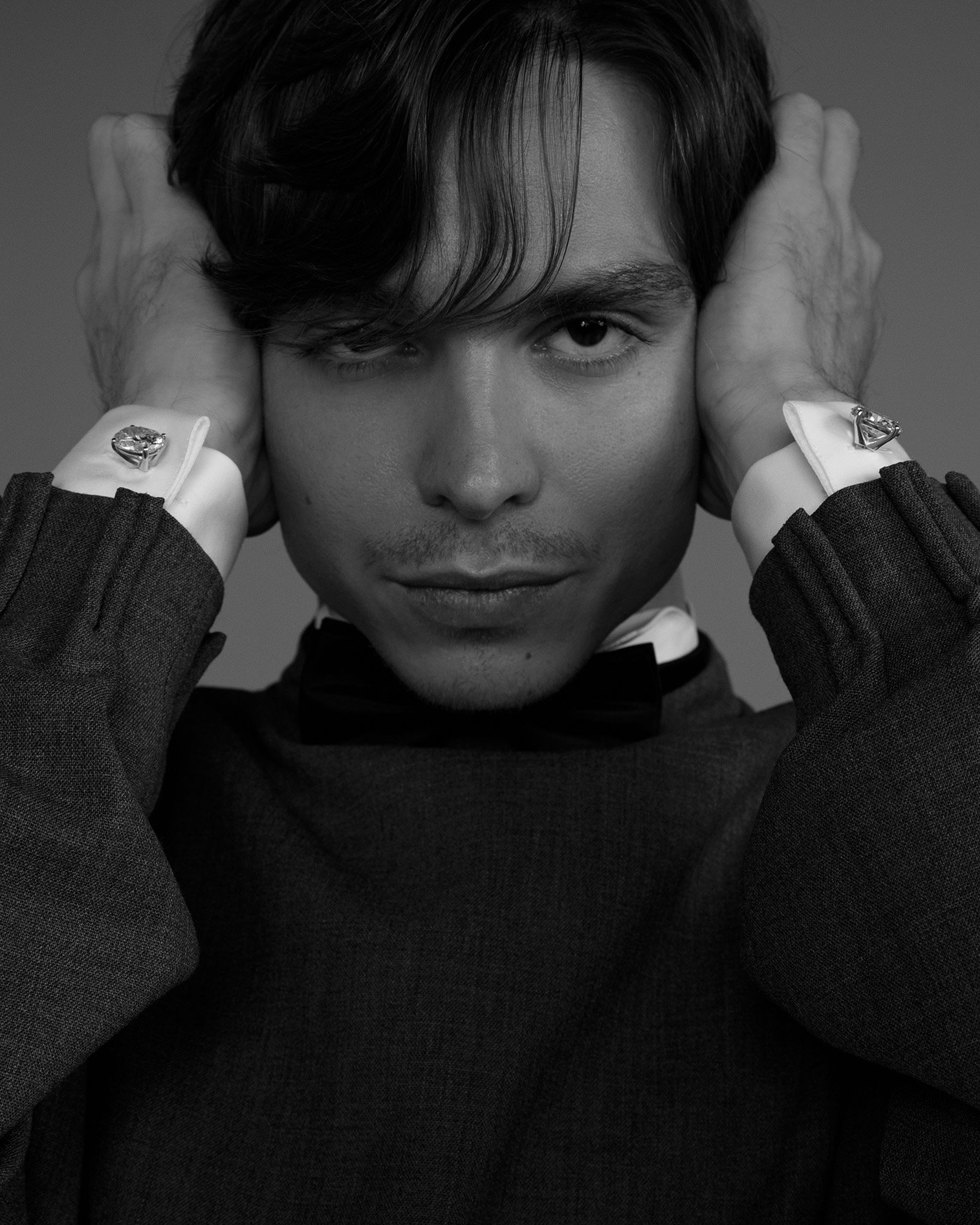 Monochrome portrait of a young adult male with hands near his face, wearing a grey sweater and cufflinks, showcasing a serious expression.
