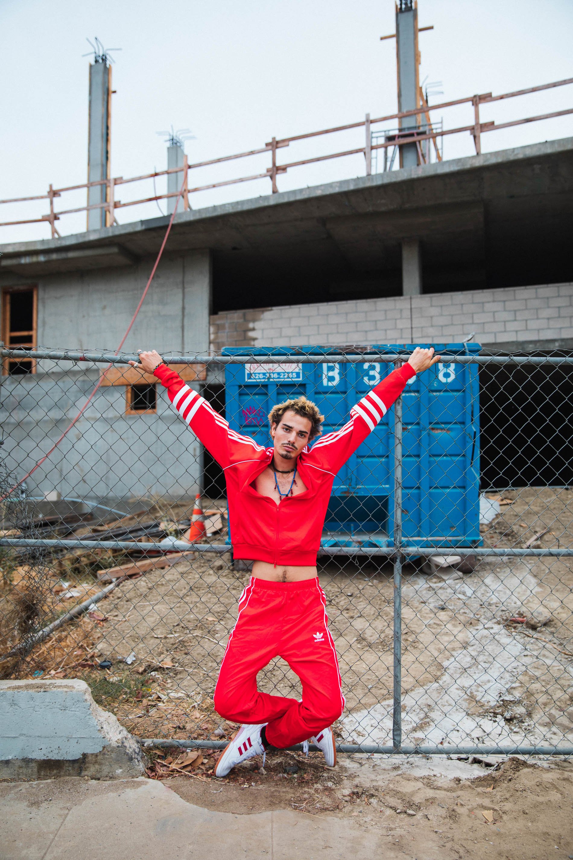 Man in red tracksuit leaps in front of a construction site with a chain-link fence, showcasing a casual and energetic pose in daylight.