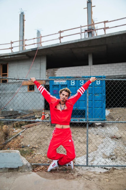 Man in red tracksuit leaps in front of a construction site with a chain-link fence, showcasing a casual and energetic pose in daylight.