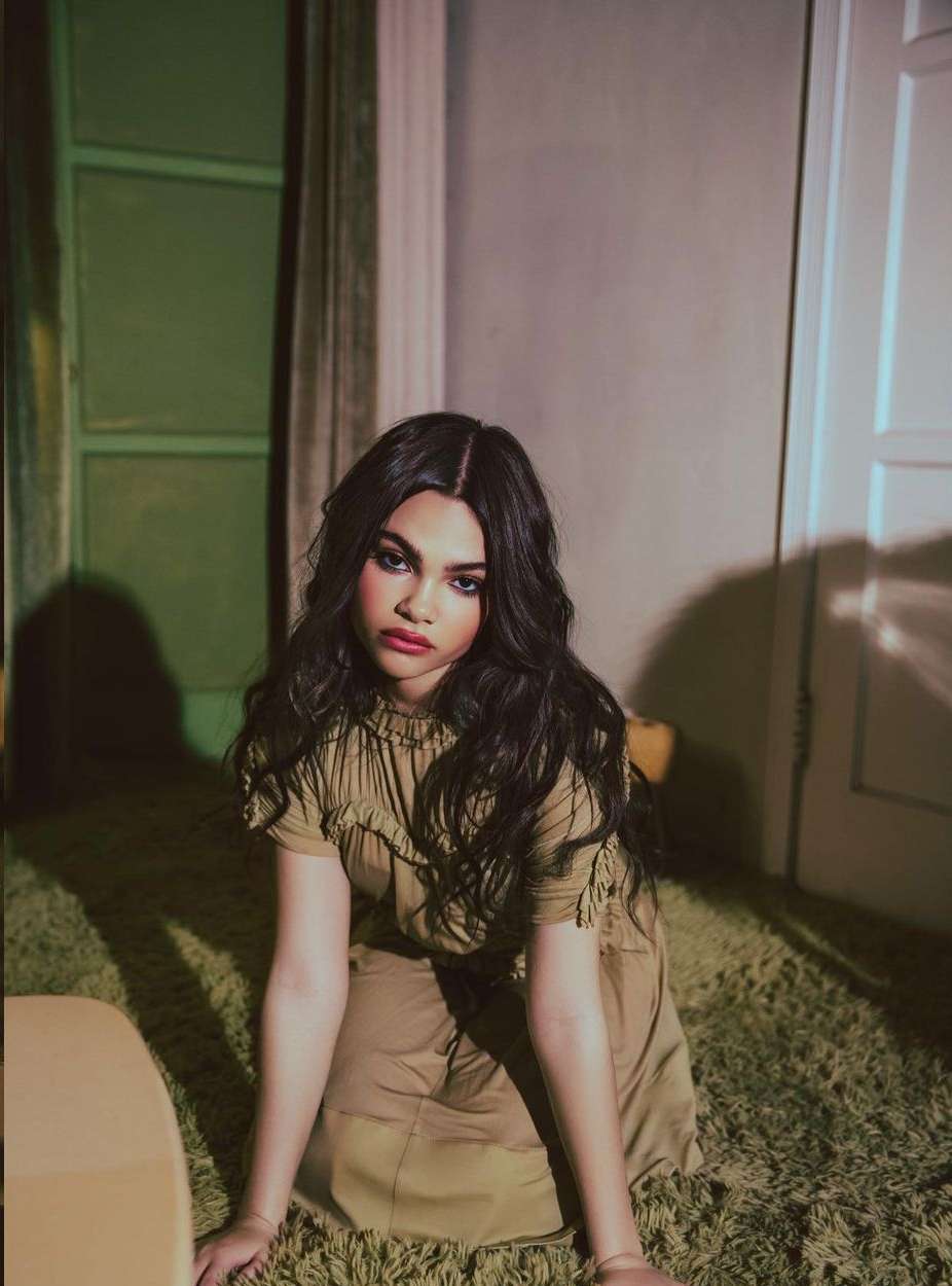 Portrait of a woman with dark hair in a light brown dress, kneeling on a green carpet. Soft lighting, interior setting, and neutral tones.