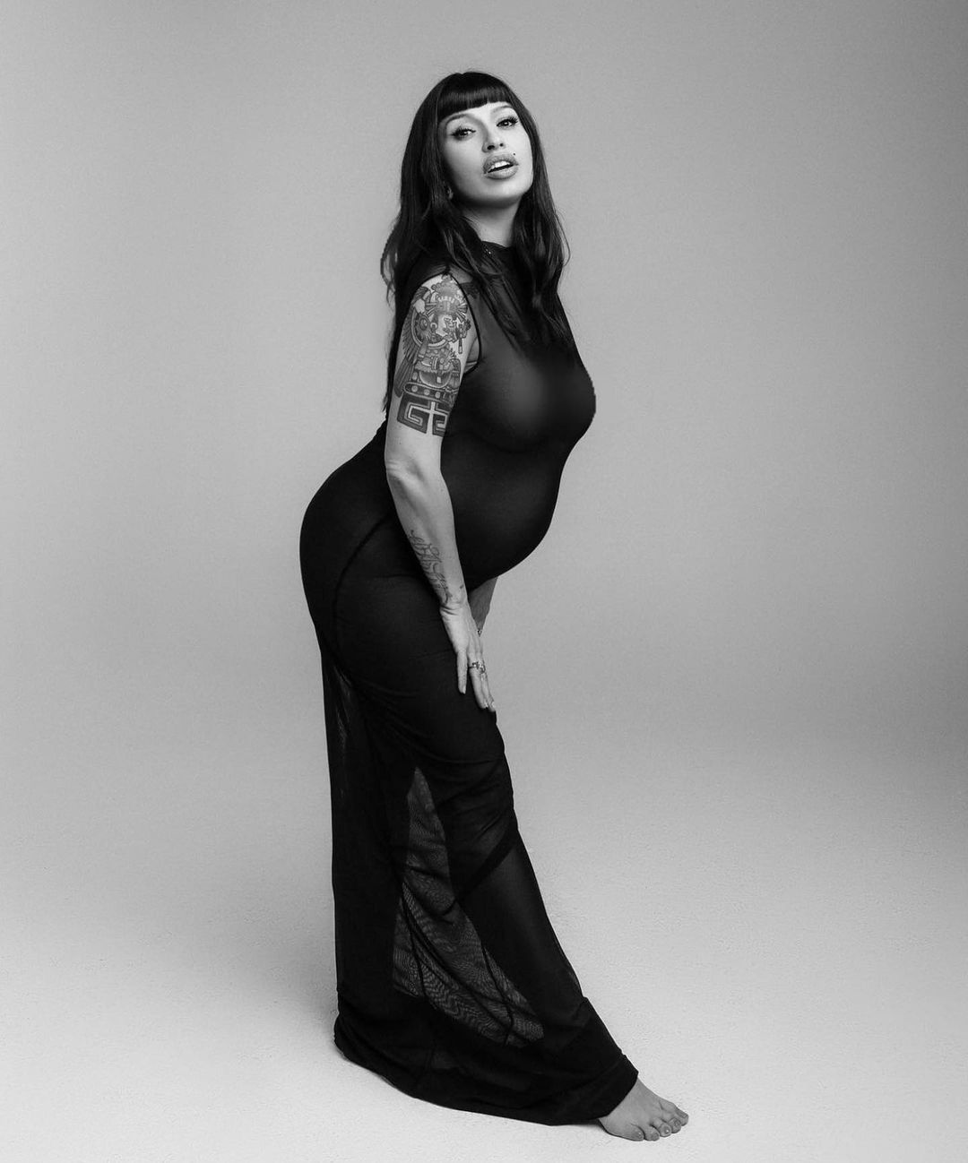 Monochrome studio portrait of a pregnant adult female with dark hair, fair skin, and tattoos, wearing a sheer black dress.