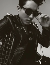 Man wearing sunglasses and a black leather jacket leaning against a metal grid against a plain grey background