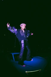 A person with dyed pink dreadlocks stands atop a blue sports car at night, wearing a patterned dark jacket and leather trousers.