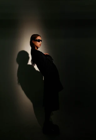A model wearing dark sunglasses and a black coat posed against a plain wall with a dramatic spotlight shadow.