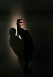 A model wearing dark sunglasses and a black coat posed against a plain wall with a dramatic spotlight shadow.