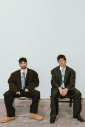 Two men in oversized dark suits and patterned ties seated on chairs against a plain white wall, photographed by Camraface.