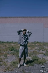 Man wearing grey sweatshirt and matching shorts posing with hands behind his head in an outdoor lot against a wall.