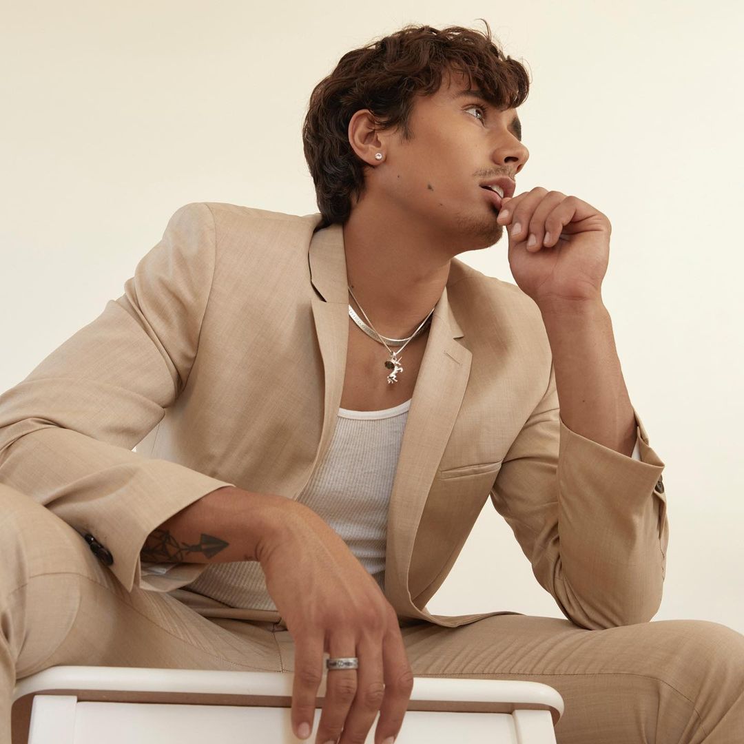 A young adult male sits in a light beige suit jacket and matching pants, wearing a white ribbed tank top and silver chain necklace.
