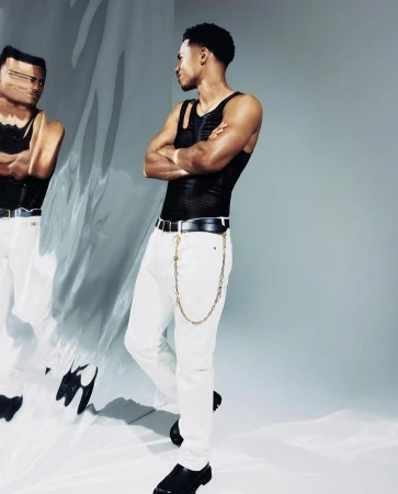 Man in black tank top and white jeans with gold chain belt standing before a reflective distorted surface by Blair Caldwell