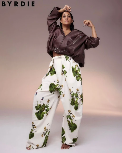 Adult woman wearing a brown leather jacket and patterned pants poses in a studio. Fashion editorial shot with neutral background.