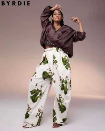 Adult woman wearing a brown leather jacket and patterned pants poses in a studio. Fashion editorial shot with neutral background.