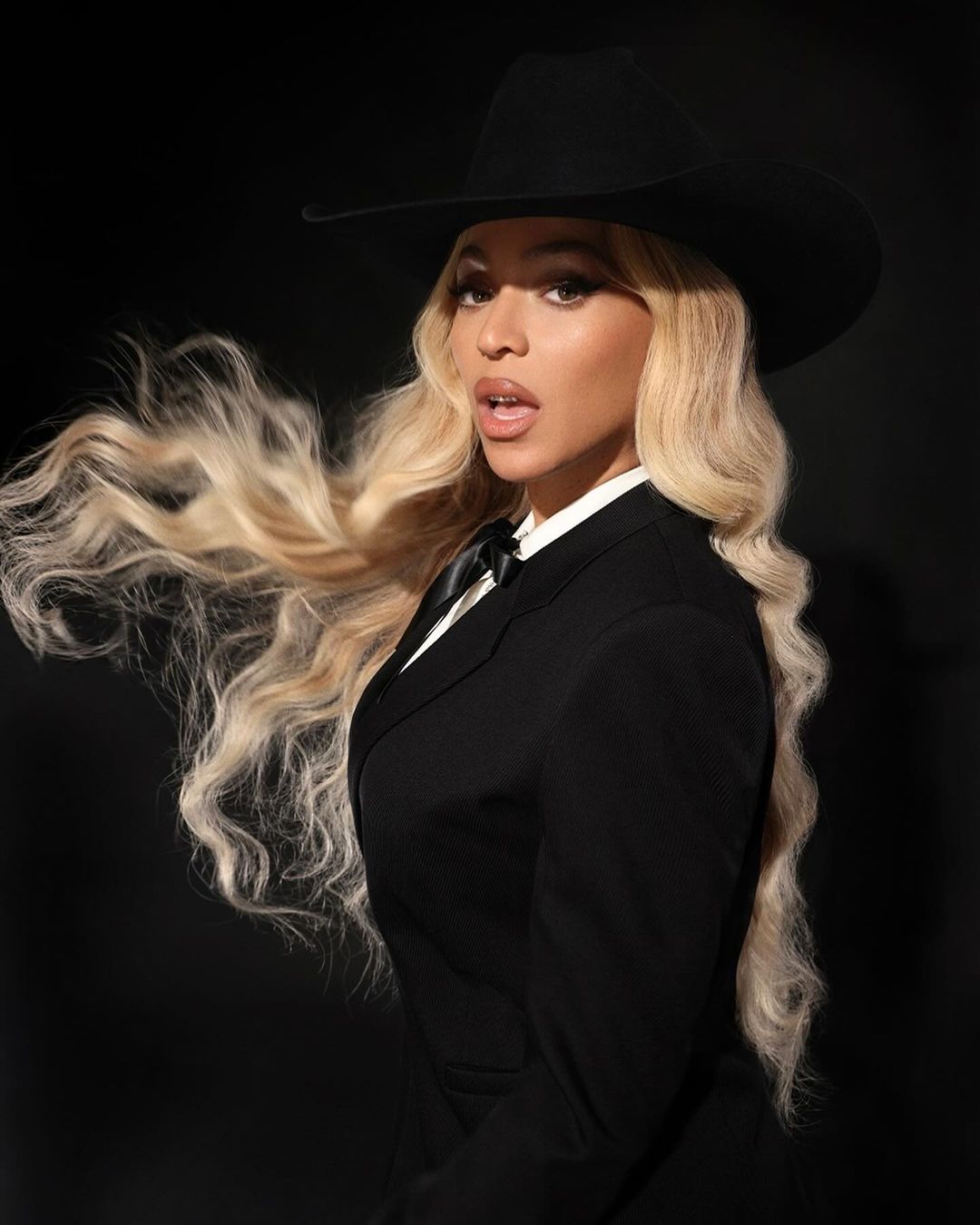 Portrait of a young adult female with blonde wavy hair wearing a black cowboy hat and black suit against a dark background.