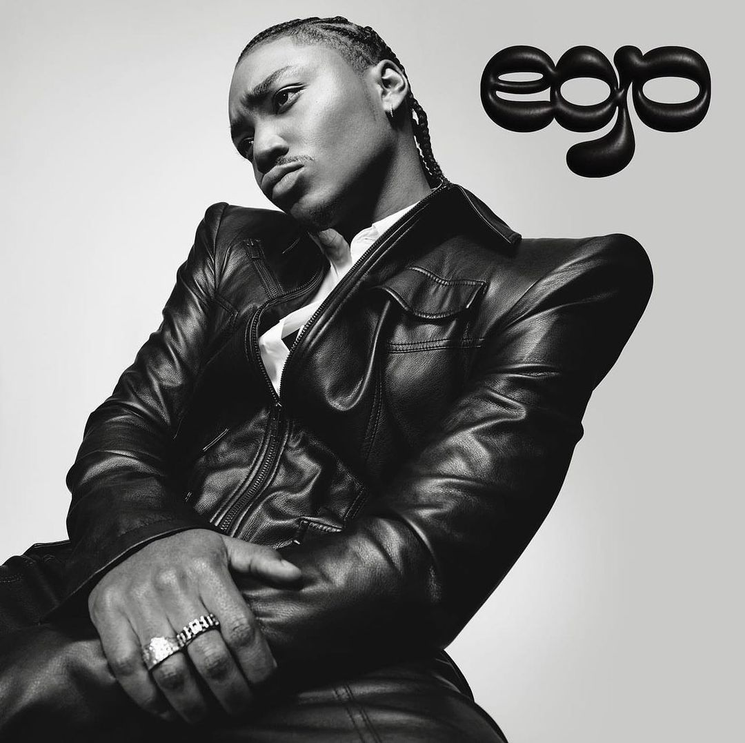 Black and white studio portrait of a young adult with cornrow braids wearing a black leather jacket with the word 'ego' visible.