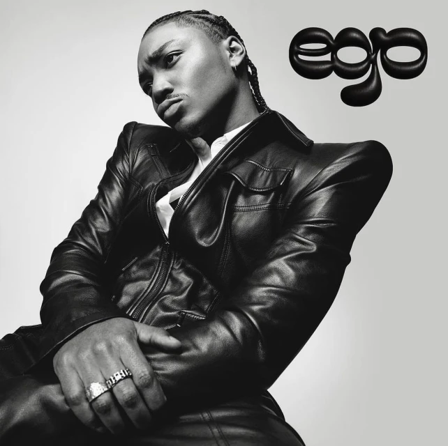 Black and white studio portrait of a young adult with cornrow braids wearing a black leather jacket with the word 'ego' visible.