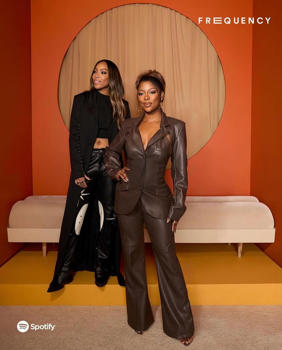 Two women pose in an indoor studio setting with orange walls and a light-beige couch. One woman wears a brown pantsuit, the other a long black coat.