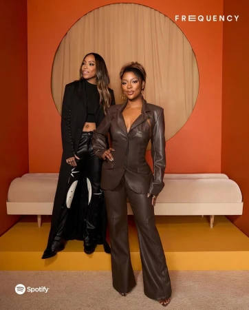 Two women pose in an indoor studio setting with orange walls and a light-beige couch. One woman wears a brown pantsuit, the other a long black coat.