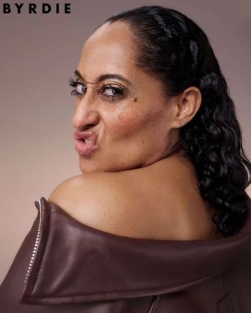 Portrait of a woman with dark curly hair and brown leather jacket looking over her shoulder making a funny face with the logo BYRDIE in corner.