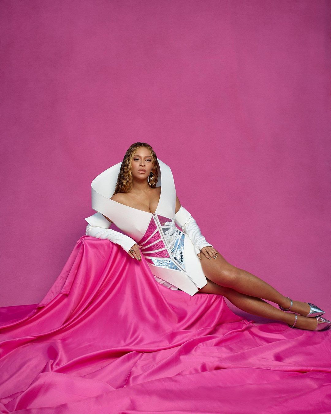 Beyonce poses in a white and silver dress with exaggerated collar on a pink draped surface against a pink backdrop in a studio shot.