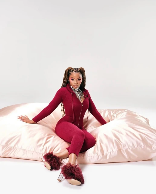 Elegant woman with long braided hair poses in a maroon jumpsuit and fuzzy slippers while seated on a light pink satin pillow in a studio.