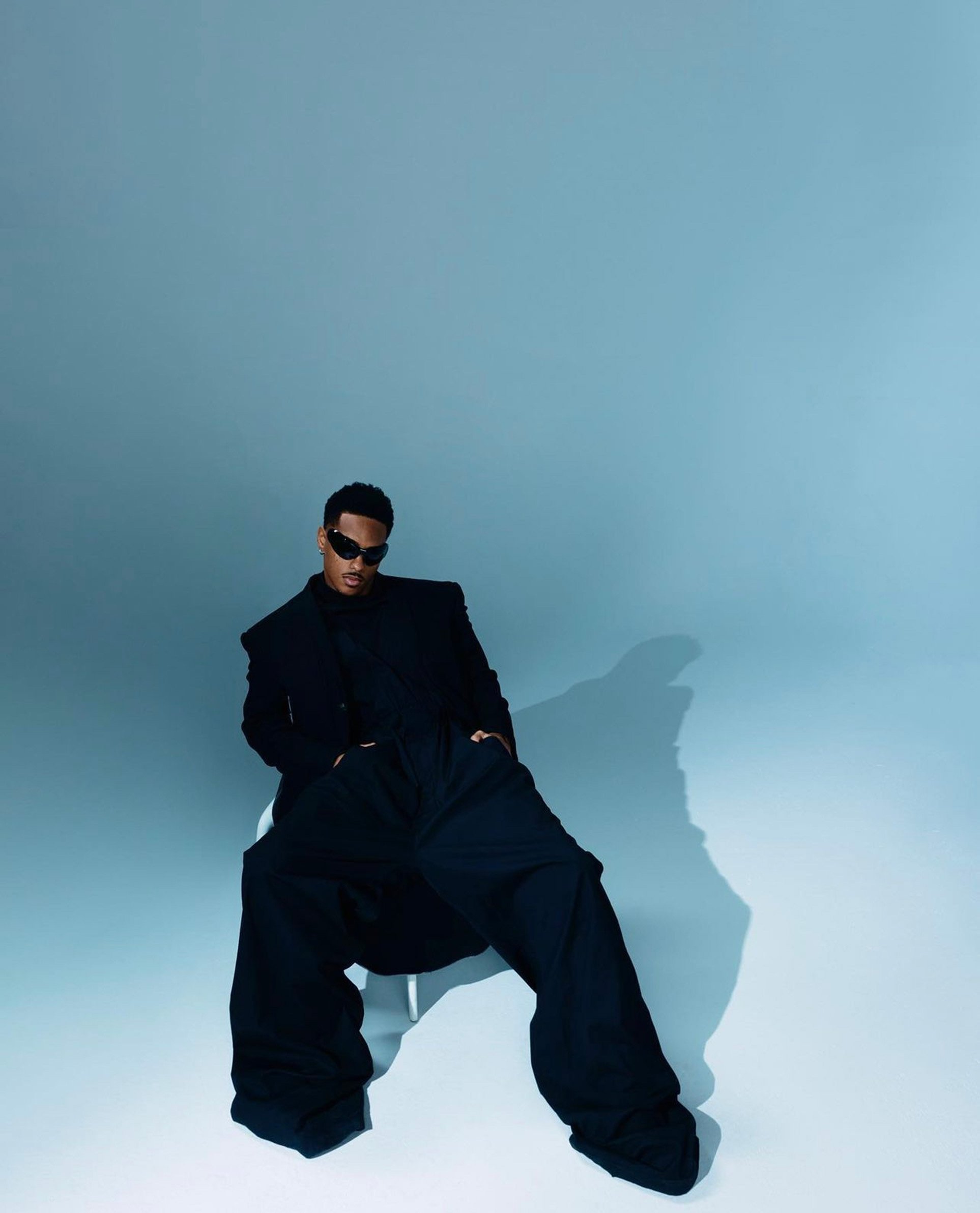 Man wearing sunglasses and a black suit seated on a chair against a light blue seamless studio backdrop by Blair Caldwell