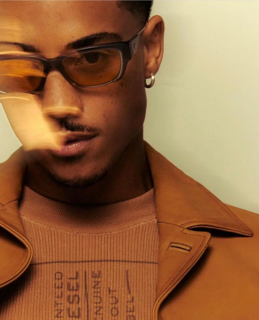 Close-up studio shot of a young adult man wearing glasses, a brown sweater and jacket, with neutral color grading and soft studio lighting.