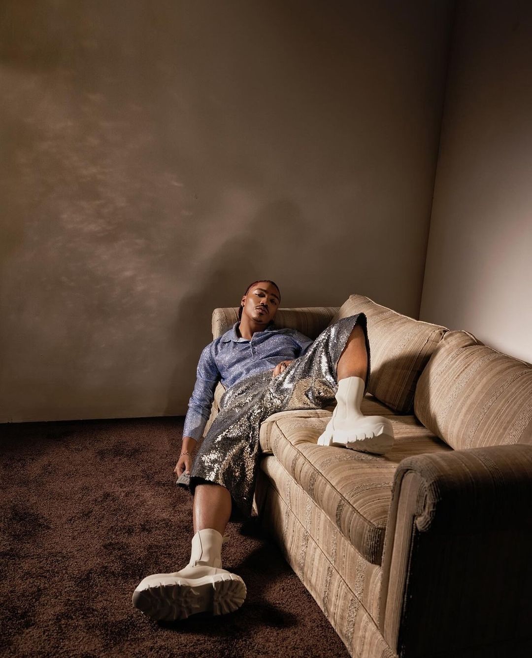 Adult male fashion model poses while reclining on a couch in a studio environment, wearing shorts, socks, and a button-down shirt.