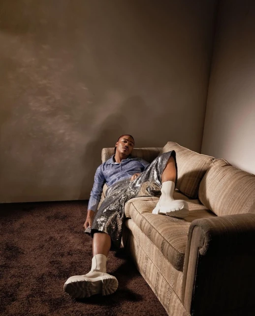 Adult male fashion model poses while reclining on a couch in a studio environment, wearing shorts, socks, and a button-down shirt.