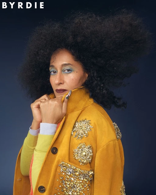 Portrait of an adult woman with an afro hairstyle wearing a yellow embellished jacket against a dark blue background.