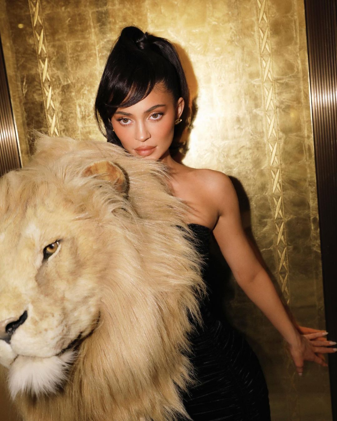 A woman poses with a lion head prop in front of a gold-patterned wall, wearing a strapless black dress with dark hair.